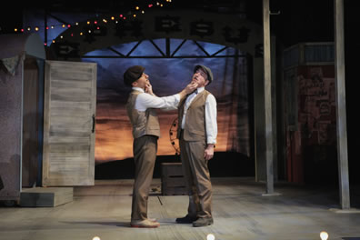 The two Dromios in brown vest and pants and white shoes, both wearing dog caps, hold each other's chins, turnng their heads to either side. In the back ground is the carnival stage, a ferris wheel silhouetted against a sunset in the backdrop, and an open door to the left.